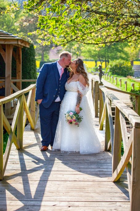 Bride and groom embrace on a wooden bridge, surrounded by greenery and sunlight.