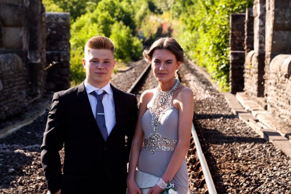 A young couple in formal attire stands on a railway track surrounded by greenery.
