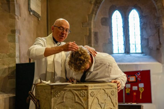 A clergyman baptising a person at a stone font inside a church.
