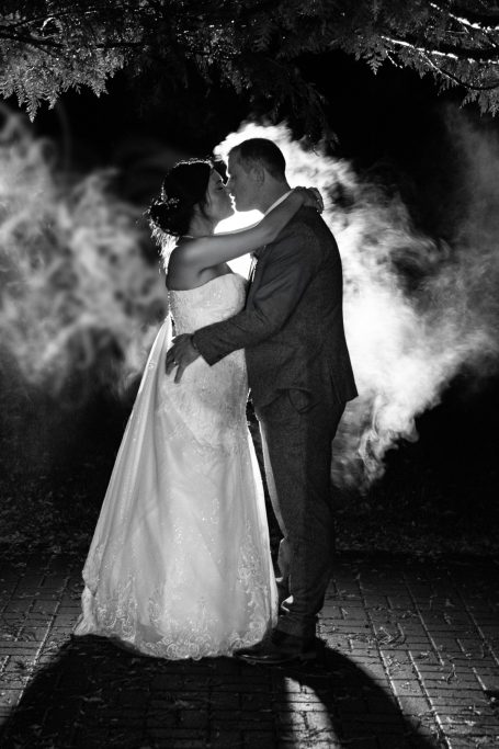 Couple in wedding attire embraces, surrounded by soft, ethereal mist.