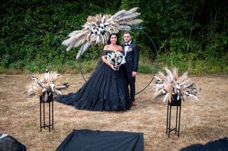 Bride in a black gown and groom in a suit, standing in a natural setting with floral arrangements.