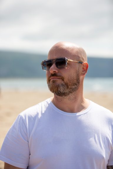 Man with a beard wearing sunglasses, standing on a beach with a distant shoreline.
