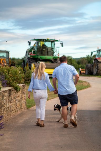 A couple walks hand-in-hand with a dog along a farm track, surrounded by tractors.