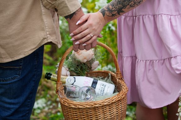 A couple holding hands beside a basket containing drinks and a teddy bear.