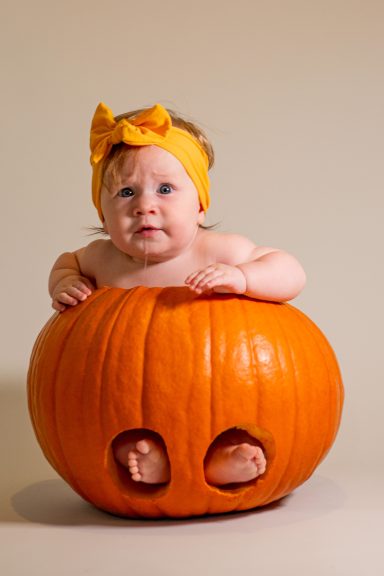A baby sitting inside a carved pumpkin, wearing a yellow bow headband.