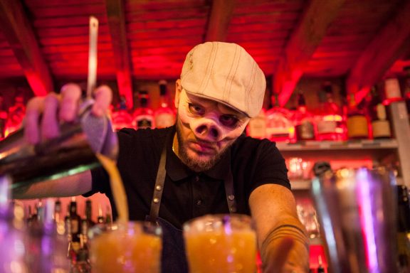 Bartender in a cap pouring cocktails at a vibrant bar with pink lighting.