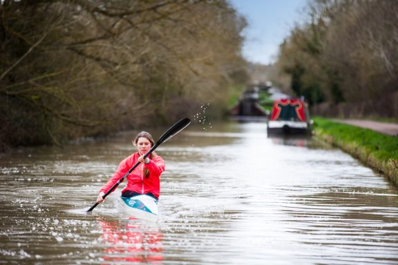 A kayaker paddles down a calm canal, with trees and boats lining the banks.