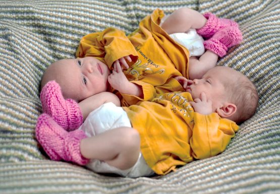 Two babies in matching orange outfits with pink booties, cuddling on a striped blanket.