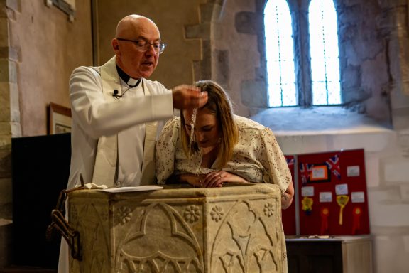 A priest baptising an adult woman at a church font. Natural light filters through stained glass windows.