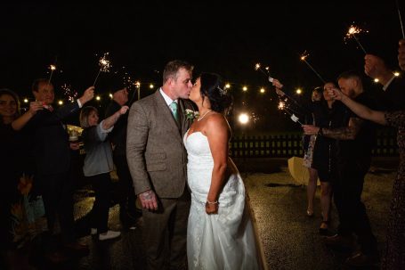 A newlywed couple kissing while surrounded by guests holding sparklers.
