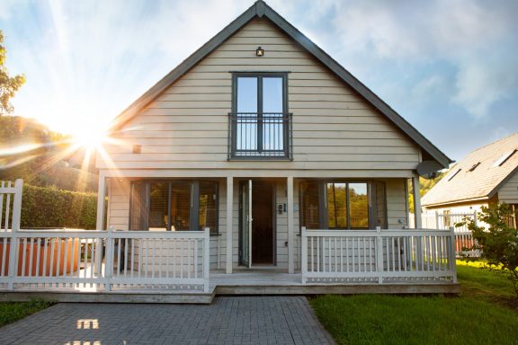 Modern two-storey cabin with a front porch and large windows, sun shining behind.