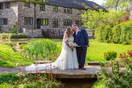 Bride and groom on a stone bridge in a lush garden outside a historic stone house.