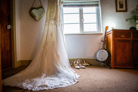Bridal dress hanging by a window with shoes and a banjo nearby.