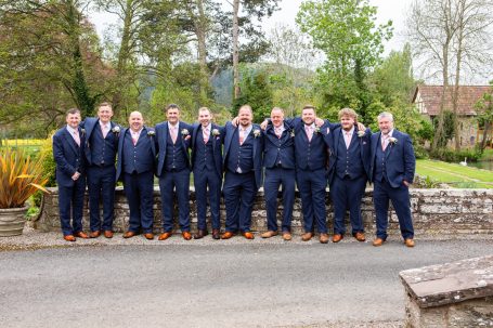 Group of 12 men wearing navy wedding suits and brown shoes, posing outdoors with greenery in the background.