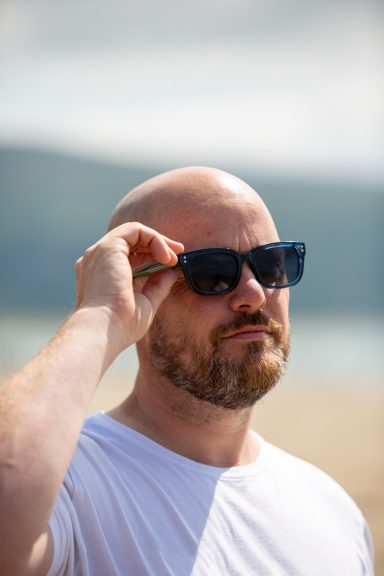 A man wearing sunglasses adjusts them while standing on a beach.