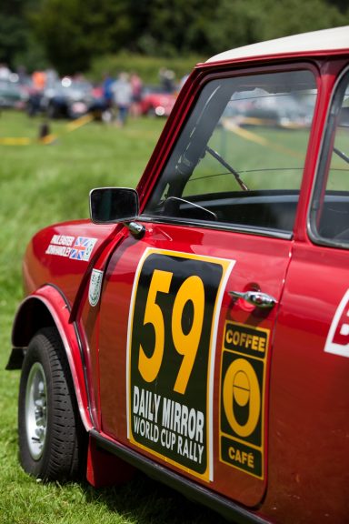 Red vintage car with racing number 59 parked in a grassy field, surrounded by distant vehicles.