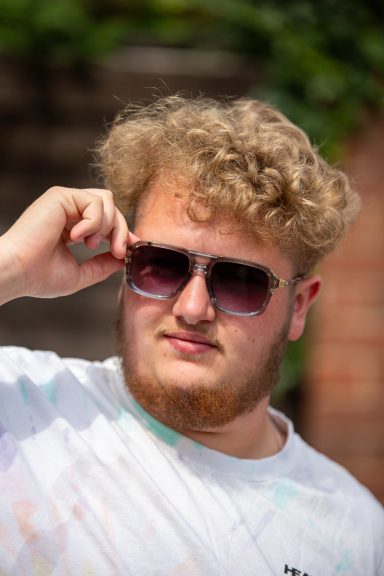 Young man adjusting sunglasses, with curly hair, wearing a white t-shirt.