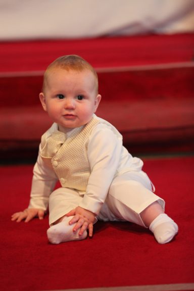 A small baby seated on a red carpet, wearing a light-coloured formal outfit.