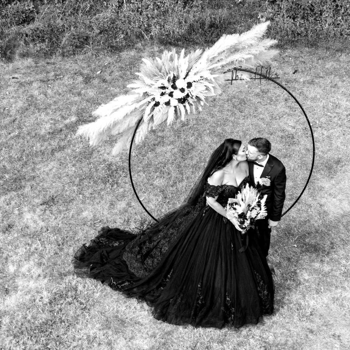 A wedding couple share a kiss under a floral arch in a grassy setting.