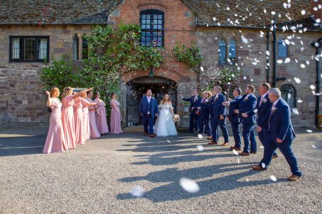 Bride and groom surrounded by guests throwing confetti outside a rustic venue.