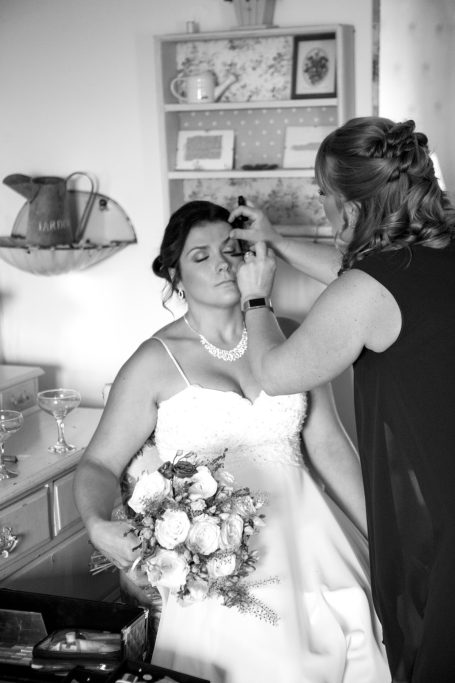 Bride receiving makeup application while holding a bouquet of flowers.