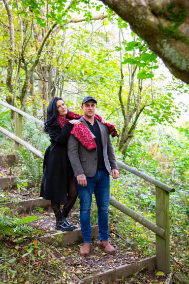 A couple stands on wooden steps in a lush green woodland setting.