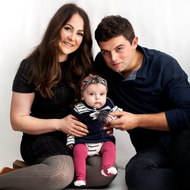 A young family sits together, smiling, with a baby in the mother's lap.