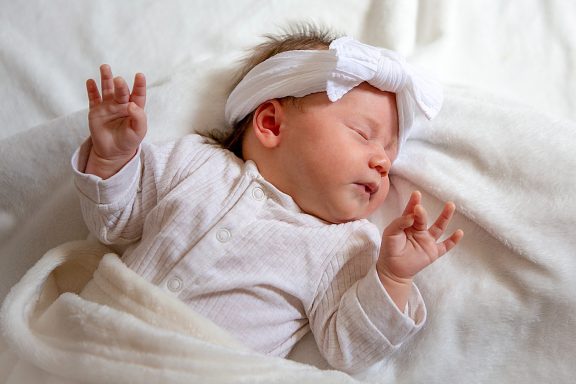 A sleeping baby in a white outfit and headband, wrapped in a soft blanket.