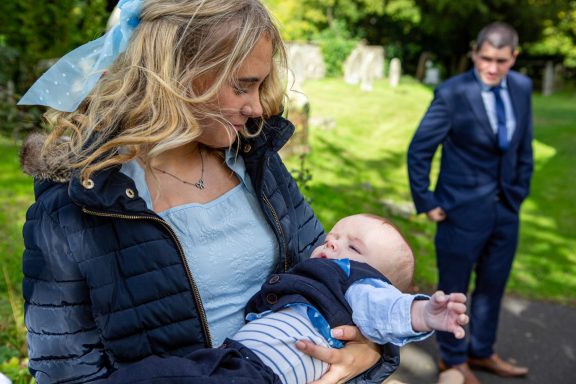 A woman holding a baby in a churchyard, while a man stands nearby in formal attire.