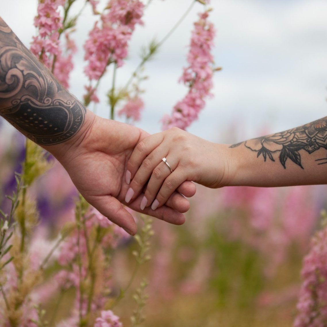 Two hands with tattoos holding each other among pink flowers.
