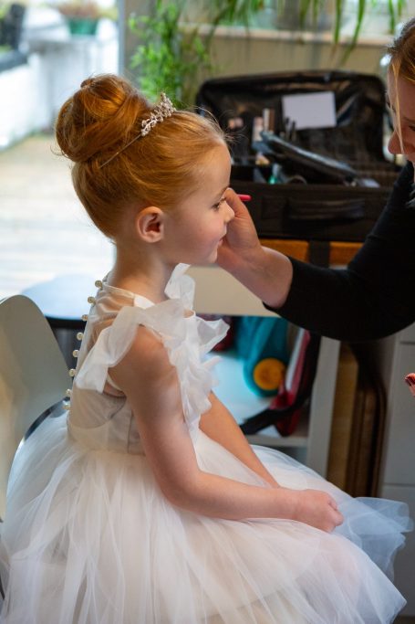 A young girl in a white dress receiving makeup, seated and smiling.