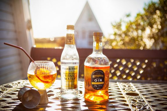 A glass of cocktail with bottles of spirits on a sunlit table outdoors.