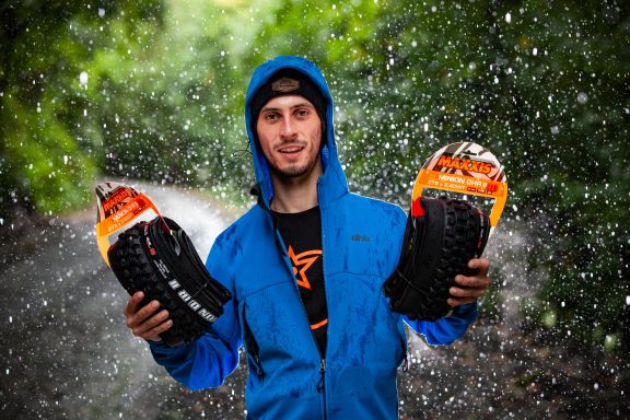 Person in a blue waterproof jacket holding two colourful packaged  mountain bike tyres in a raining forest setting.