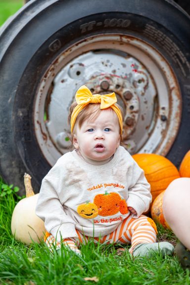 A baby wearing a pumpkin-themed outfit sits among pumpkins near a tractor tire.