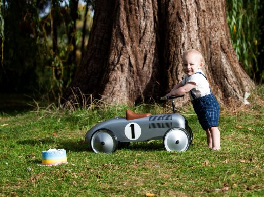 Baby pushing a vintage toy car in front of a large tree in a grassy area.