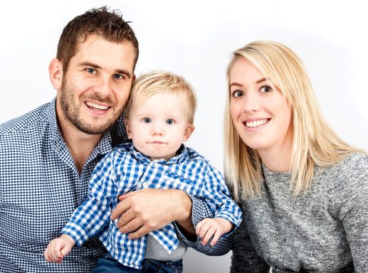 A smiling couple holding a baby, all dressed casually against a white background.