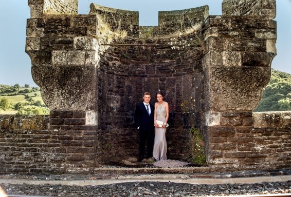 A couple stands together in formal attire against a stone castle backdrop.