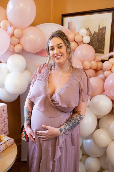Pregnant woman in a pink dress, smiling, surrounded by pink and white balloons.