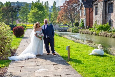 Bride and groom stand by a serene canal, with a swan nearby and greenery surrounding.