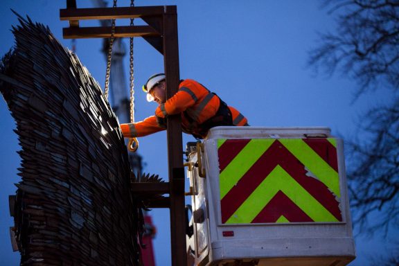 Worker in high-visibility clothing using tools from a raised platform at twilight.