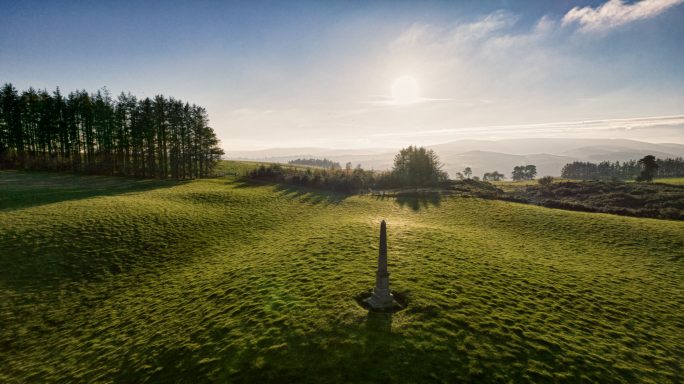 A peaceful landscape with rolling hills, trees, and a stone marker under a clear sky.