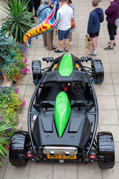 A black and green sports car with a sleek design, surrounded by tropical plants, people and a helium balloon with the words "Hot Wheels".