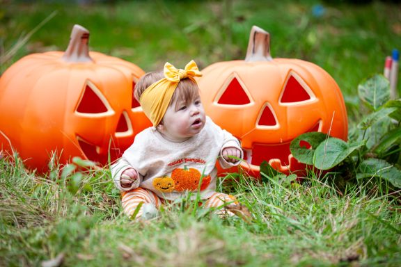 Baby sitting among carved pumpkins, wearing a yellow headband and a costume.