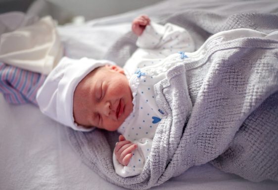 Newborn baby wrapped in a grey blanket, wearing a white hat and sleeping peacefully.