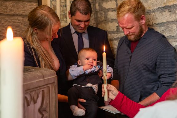 A family gathers for a baptism, holding a baby near a lit candle.