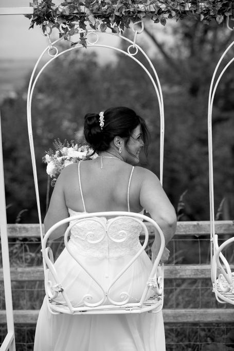 Bride in a wedding dress sitting on a white swing, facing away, surrounded by nature.