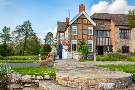 A bride and groom stand in front of a traditional house, surrounded by greenery.