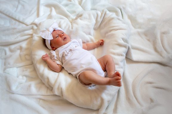 A sleeping newborn in a white outfit and headband, lying on a fluffy heart-shaped cushion.