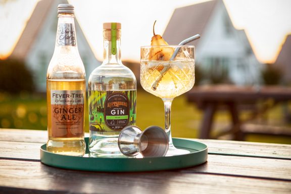 A cocktail with a pear garnish, two bottles, and a jigger on a wooden table outdoors.