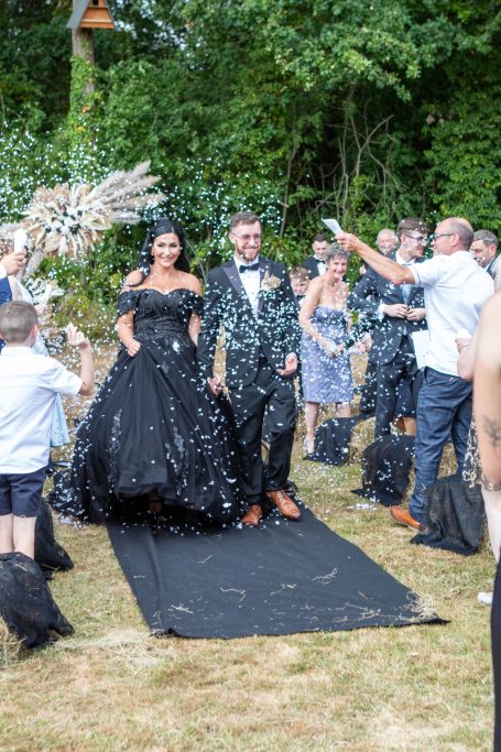 A couple in formal attire walks down an outdoor aisle, surrounded by guests and greenery.